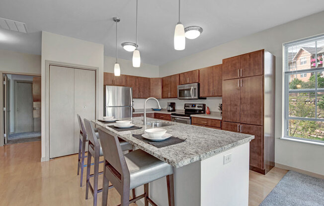 A spacious kitchen here at Vue at Pinnacle Park with warm wood cabinets, granite countertops, stainless steel appliances, and a large island with bar seating, pendant lighting, and a bright window for natural light.