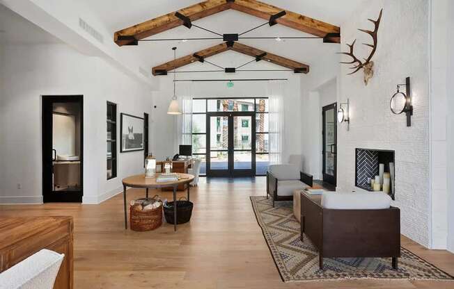 A welcoming community lounge here at Reflect at Dobson Ranch featuring vaulted ceilings with exposed wood beams, warm wood flooring, a round accent table with decorative baskets, a cozy seating area beside a white brick fireplace with wall sconces, and glass doors that open to outdoor views, creating an inviting gathering space.