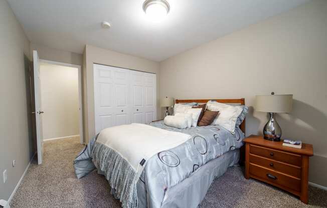 Bedroom with plush carpeting, bed with blue bedding, and wooded nightstand with lamp on top and a large white closet.