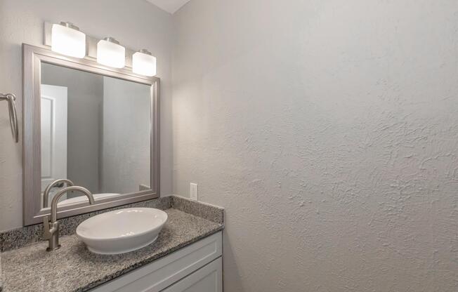 A modern bathroom featuring a light gray wall, a sleek gray countertop with a white vessel sink, and a decorative mirror above. Three light fixtures provide illumination above the mirror, enhancing the contemporary design of the space.