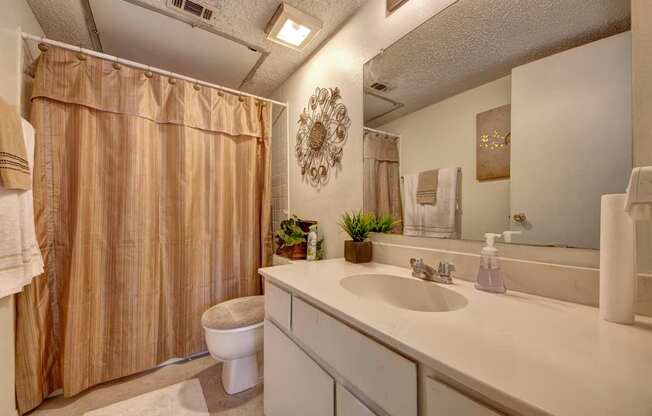 A clean bathroom here at Summertree Place featuring a white vanity with integrated sink, a large wall mirror, neutral countertop, striped shower curtain, tiled flooring, and neatly arranged towels, creating a bright and functional space with simple decorative accents.