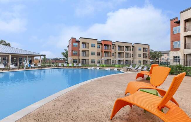A swimming pool with orange chairs and apartment buildings in the background.