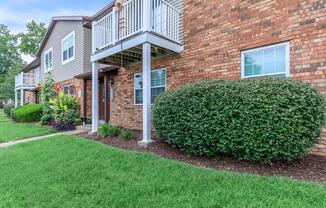 A well-manicured apartment community with a brick exterior. The front features two balconies, neatly trimmed hedges, and colorful landscaping. The lawn is green and lush, contributing to a tidy and inviting atmosphere.