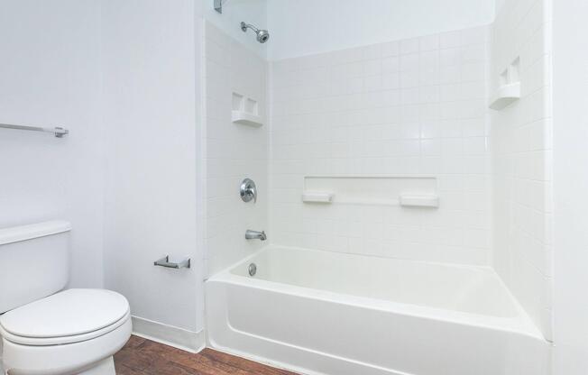 A clean and modern bathroom featuring a white bathtub with a showerhead, a toilet, and light-colored walls. The flooring is wood-like, adding warmth to the space. There are built-in shelves in the bathtub area for convenience.