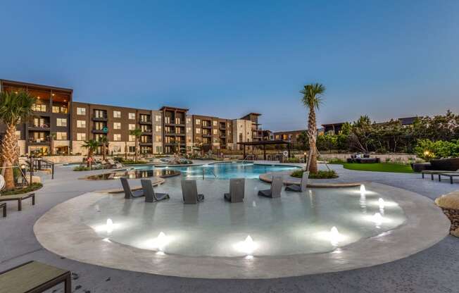 A round pool with lights in the middle surrounded by chairs and a building in the background.
