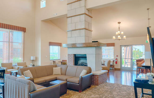 A welcoming community lounge here at Dakota Ridge featuring a large stone fireplace with brick accents, cozy sectional seating with neutral cushions, wood flooring, tall windows that bring in natural light, soft area rugs, and nearby tables and chairs that create a comfortable space for relaxing or gathering with neighbors.