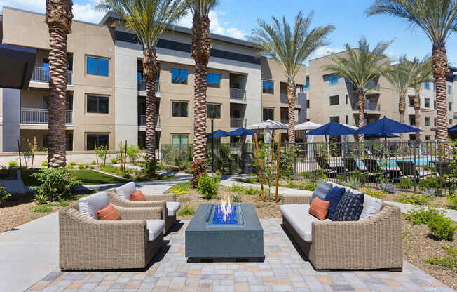 A landscaped outdoor seating area here at Reflect at Dobson Ranch featuring woven lounge chairs with neutral cushions arranged around a sleek rectangular fire feature, surrounded by palm trees, flowering greenery, and paved walkways, with modern apartment buildings and a resort-style pool area visible in the background under clear blue skies.