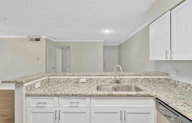 A modern kitchen with granite countertops, a stainless steel sink, and white cabinetry. The kitchen opens to a light-colored living area featuring a textured ceiling and neutral walls. An entryway is visible in the background, along with appliances integrated into the cabinetry. Bright and spacious design.