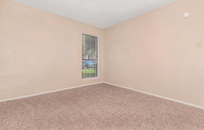 Empty room with beige walls and light brown carpeting. A single window with vertical blinds allows natural light to enter, revealing a view of greenery outside. The space is unfurnished, creating a blank canvas suitable for various uses.
