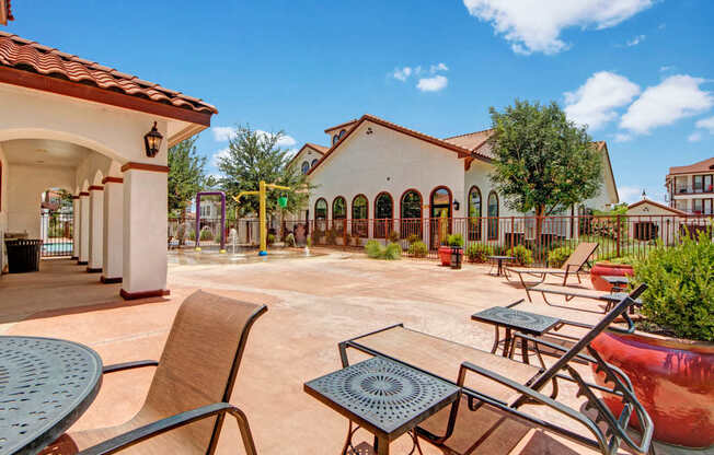A relaxing outdoor seating area here at Andalucia Villas with comfortable lounge chairs and tables on a spacious patio, overlooking the splash pad and surrounded by Spanish-style architecture, mature trees, and bright blue skies for a laid-back, resort-inspired feel.