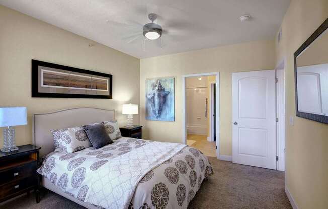 A serene bedroom here at Tuscany at Faudree with plush carpet, soft neutral walls, and a comfortable bed dressed in patterned bedding. Matching nightstands with table lamps flank the bed, while framed artwork and a ceiling fan add style. An open doorway leads to an attached bathroom, creating a calm and inviting retreat.