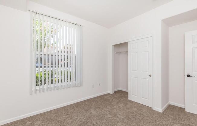 An empty room with light-colored walls and carpet. A large window covered by vertical blinds allows natural light to enter, and there is a closet with double doors on one side. The overall atmosphere is bright and spacious, with a modern, clean design.