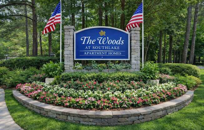A sign for The Woods at Southlake Apartment Homes is surrounded by flowers and American flags.