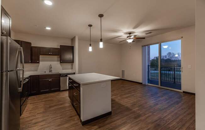 A modern kitchen and living area here at The Park featuring dark cabinetry, a bright island, warm wood floors, pendant lights, and sliding glass doors that bring in natural light and create an open, inviting feel.