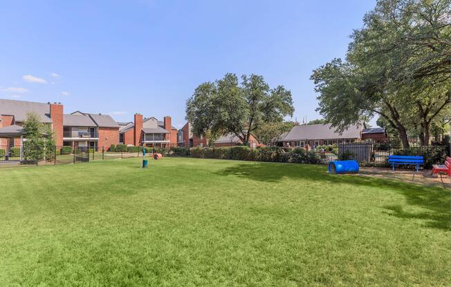 A spacious green park area surrounded by residential buildings, featuring trees, a blue tunnel structure, a red bench, and a fenced perimeter. The sky is clear with a few clouds, creating a bright and inviting atmosphere.
