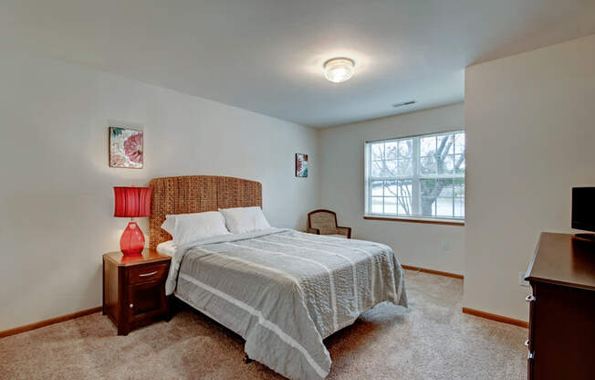 A comfortable bedroom here at Arbor Ridge featuring a woven headboard bed, soft neutral bedding, and warm wood furniture. A red bedside lamp and bright window add cozy charm and natural light.