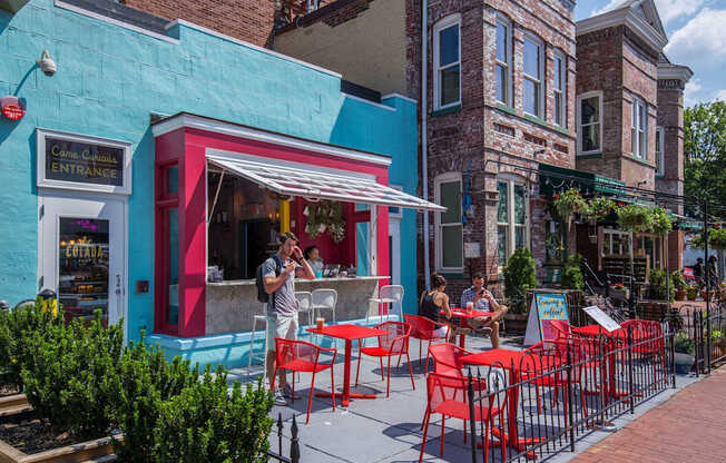 A man is standing outside a restaurant with red chairs and tables.