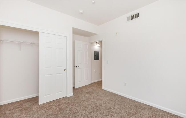 A small, empty room featuring beige carpet, white walls, and a closet with a sliding door. There are two doors: one leading to another room and another door that appears to lead to a bathroom or storage. The ceiling is plain with a couple of overhead lights. The space is well-lit and uncluttered.