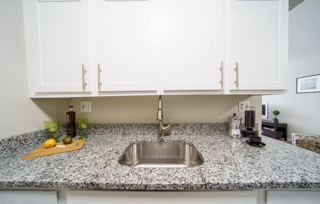 Stainless steel sink with white upper cabinets and  grey and white stone counters.