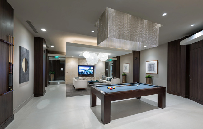 A clubhouse with dark wood-paneled walls, a billiards table under a rectangular chandelier with hanging silver chains, and two long white couches in front of a large HDTV.
