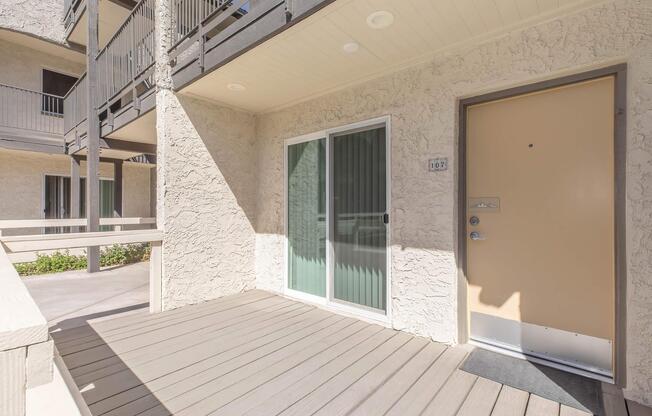 Exterior view of an apartment entrance featuring a beige wall and a wooden deck. The door is light brown with a silver handle, and there are large windows with green vertical blinds beside it. The surrounding area includes a walkway and additional apartment balconies visible in the background.