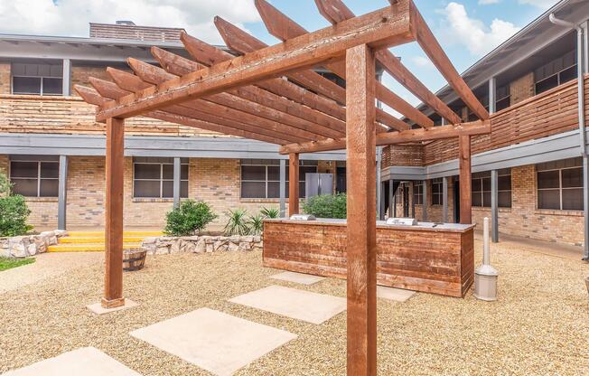 A rustic wooden pergola with slatted roofing stands in a courtyard, featuring a wooden service counter and stone paths. Surrounding the area are low-maintenance plants and gravel, complemented by an inviting atmosphere of a contemporary building with brick walls and large windows.