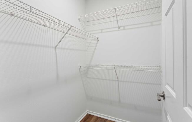 Empty closet with white wire shelving on two walls, light-colored walls, and hardwood flooring. The door is partially visible on the right side. Shadows from the shelves create a pattern on the wall.
