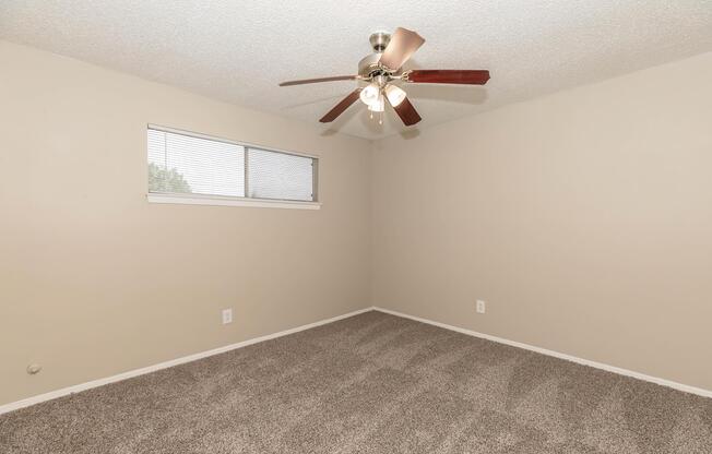 Empty room featuring beige walls and a light brown carpet. The room includes a ceiling fan with four lights and a small window with blinds, allowing natural light to enter. The space is clean and unfurnished, providing a blank canvas for decoration.