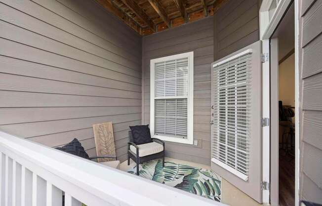A balcony with a white railing, a window with white blinds, and a chair with a black cushion.