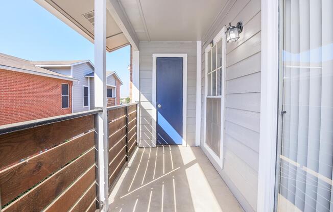 A view of a balcony with a blue door and wooden railing. Sunlight casts shadows on the concrete floor, highlighting the wooden planks of the railing. In the background, there are several residential buildings, including brick and light-colored houses, under a clear blue sky.