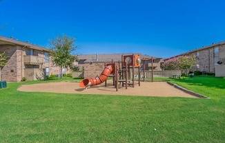 A playground with a red slide in the middle of a grassy area.