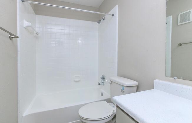 A clean, empty bathroom featuring a white bathtub with a shower curtain, a toilet, and a sink with a countertop. The walls are light gray, and there is a mirror above the sink. The space appears modern and well-maintained.