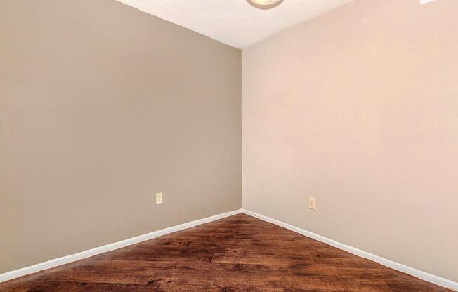 A sparsely furnished room featuring a corner with beige walls and a brown wooden floor. There is a ceiling light fixture, and the space appears empty and devoid of furnishings or decorations, creating a minimalist atmosphere.