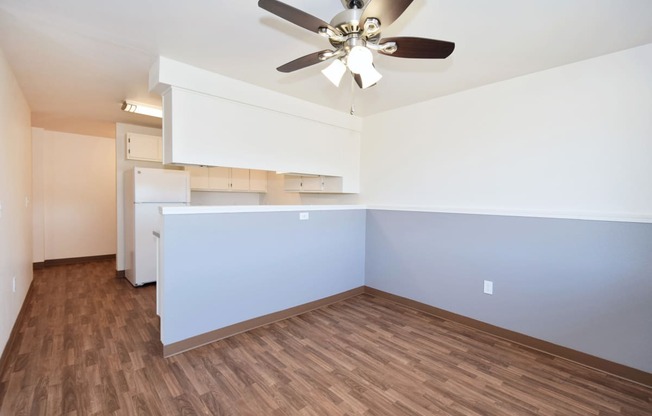 an empty living room with a ceiling fan and a kitchen in the background