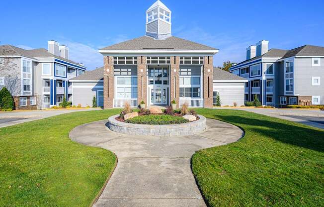 A landscaped exterior clubhouse view here at Retreat at McAlpine Creek featuring a central brick-and-siding building with tall windows, covered entry, and a cupola roof detail. A curved concrete walkway leads through green lawn and a raised stone planter with shrubs, while surrounding apartment buildings frame the welcoming community focal point.