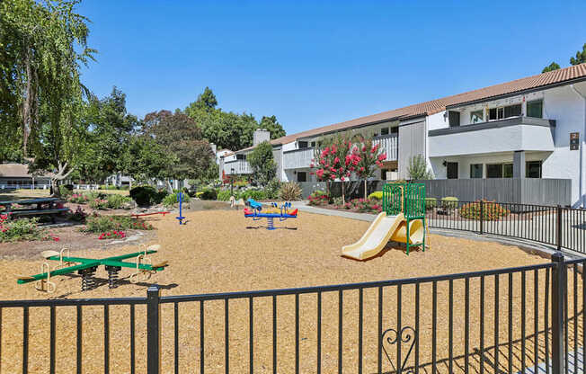 A playground with a slide, swings, and a seesaw in front of a building.