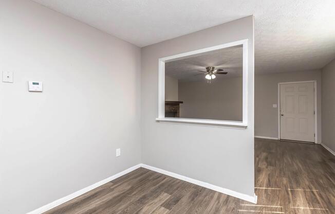 A simple interior space featuring light gray walls, a window-style opening to another room, and a ceiling fan. The floor is made of light-colored laminate, and there is a door visible on the right side. The room appears bright and inviting, with a minimalist design.