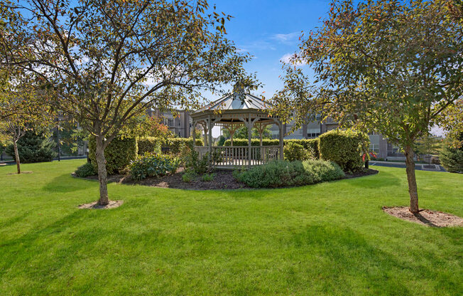 A peaceful outdoor courtyard here at Swan Creek with charming gazebo, lush green lawn, mature trees, and landscaped greenery, creating a scenic, relaxing, and inviting community space.