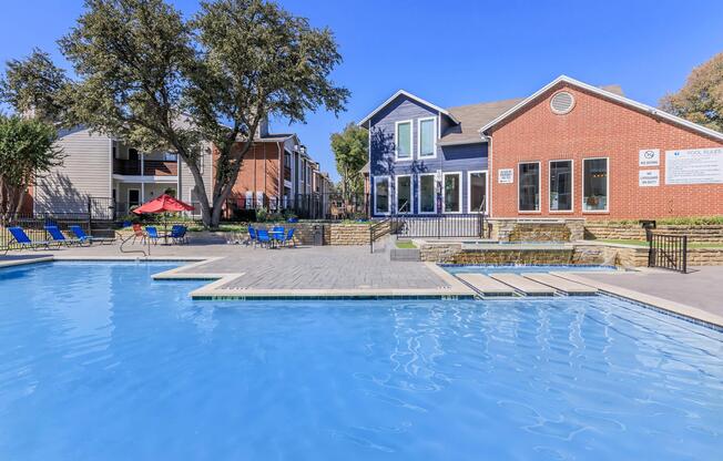 A bright blue swimming pool with lounge chairs surrounding it, in front of a modern building with a mix of brick and siding. The area is sunny, with trees nearby, and neighboring apartment buildings visible in the background. A shaded seating area with a red umbrella adds to the inviting atmosphere.