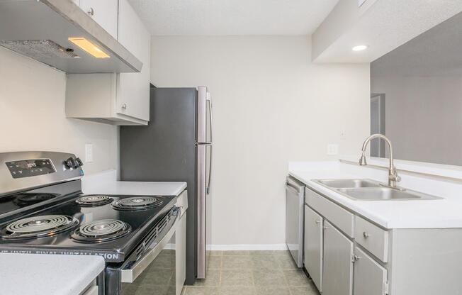 Modern kitchen with a black electric stove, stainless steel refrigerator, and double sink. The countertops are light-colored, and the cabinets are a neutral shade, creating a clean and spacious look. The floor is tiled in a light gray color, enhancing the kitchen's overall brightness.