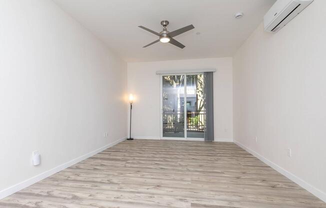 A spacious and bright living area with light wood flooring, featuring a modern ceiling fan and a standing lamp. A large sliding glass door opens to a balcony or patio area with views of greenery outside, enhancing the natural light in the room. The walls are painted in a soft, neutral color.