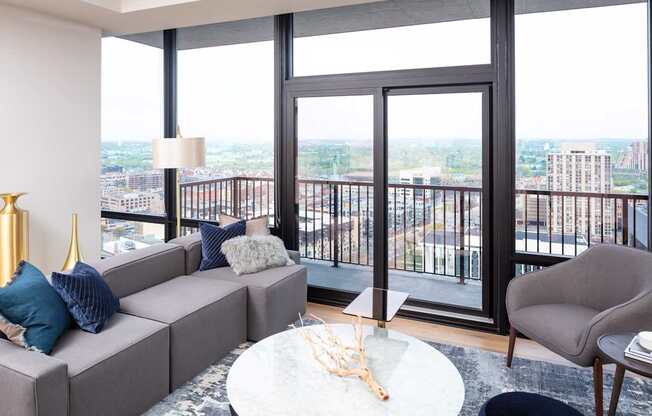 A grey couch with blue pillows sits in front of a large window with a view of the city.
