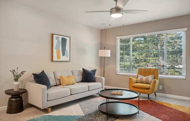 Two-Bedroom Apartments in Fremont, CA - Northgate Savoy - Furnished Living Room with Carpeted Flooring, Cream Couch, Yellow Armchair, Wooden Side Table, Standing Lamp, Round Brown Coffee Table, Large Window, Ceiling Fan, and Wall Art.