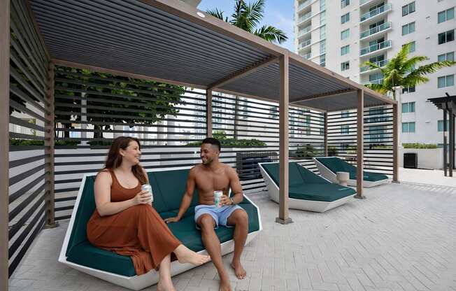 A man and a woman are sitting on a bench under a shaded area.