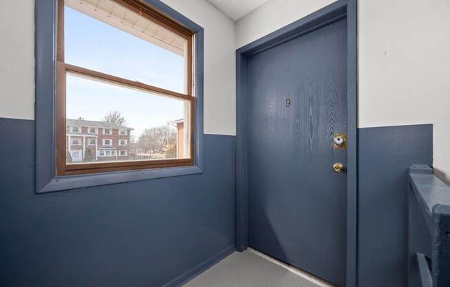 A room with a blue door and a window showing a view of a building outside.
