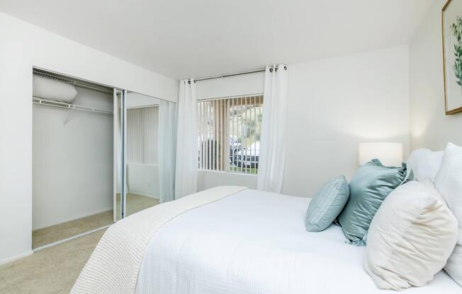 A cozy bedroom featuring a neatly made bed with light-colored bedding and decorative pillows. There's a window with sheer curtains allowing natural light, a closet with mirrored doors, and soft carpeted flooring, creating a welcoming and serene atmosphere.