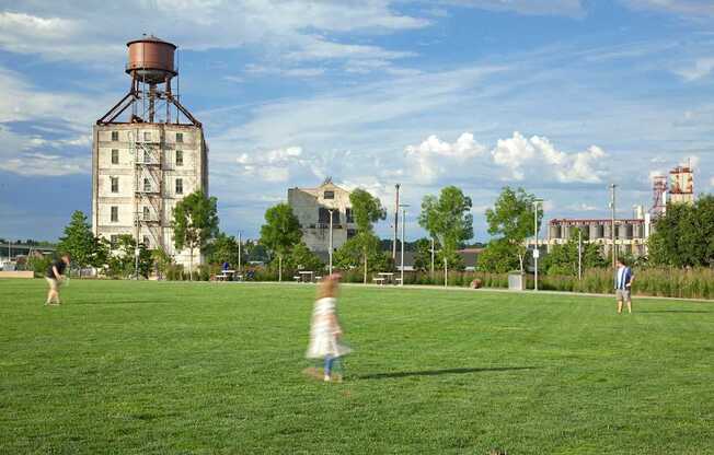 A person in a white dress is running across a grassy field with a large grain silo in the background.