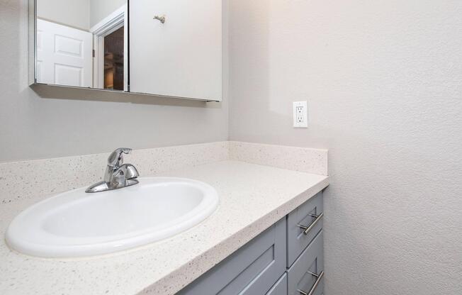 A modern bathroom sink area featuring a white circular sink set into a light-colored countertop. Below the sink are gray cabinets with metal handles. A mirror is mounted on the wall above the sink, and there is a light switch visible on the wall. The walls are painted in a neutral tone.