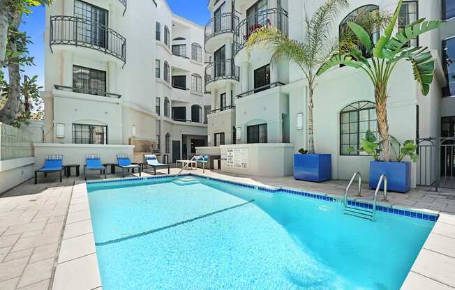 A large swimming pool in front of a white building with blue chairs around it.