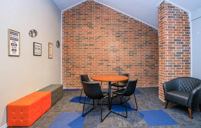 A modern meeting space featuring a round wooden table surrounded by black chairs. The floor has blue and gray tiles, and one side of the wall is adorned with a textured brick pattern. There are two decorative pieces on the wall, adding a stylish touch to the room.